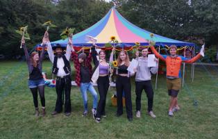 Gruppenleiter*innenschulung Eine Gruppe aus sieben jungen Menschen steht vor einem bunten Pavillion auf einer Wiese. Sie recken Sonnenblumen und ihre Juleica-Zertifikate in die Luft